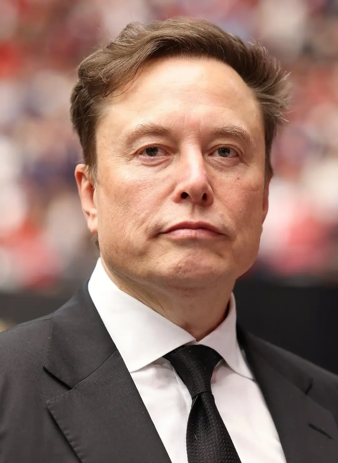 Elon Musk in a dark suit at an indoor event, photographed front-on with his hair brushed back and the front hairline visible.