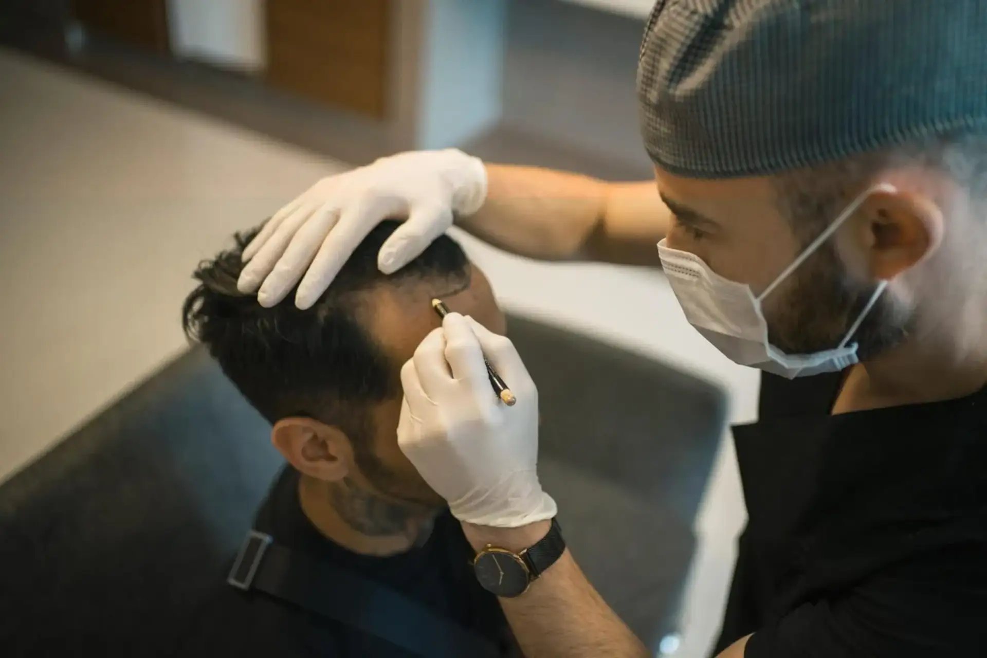 Medico con mascherina e guanti che traccia la nuova linea frontale dei capelli sulla fronte di un uomo seduto, durante la visita pre-operatoria