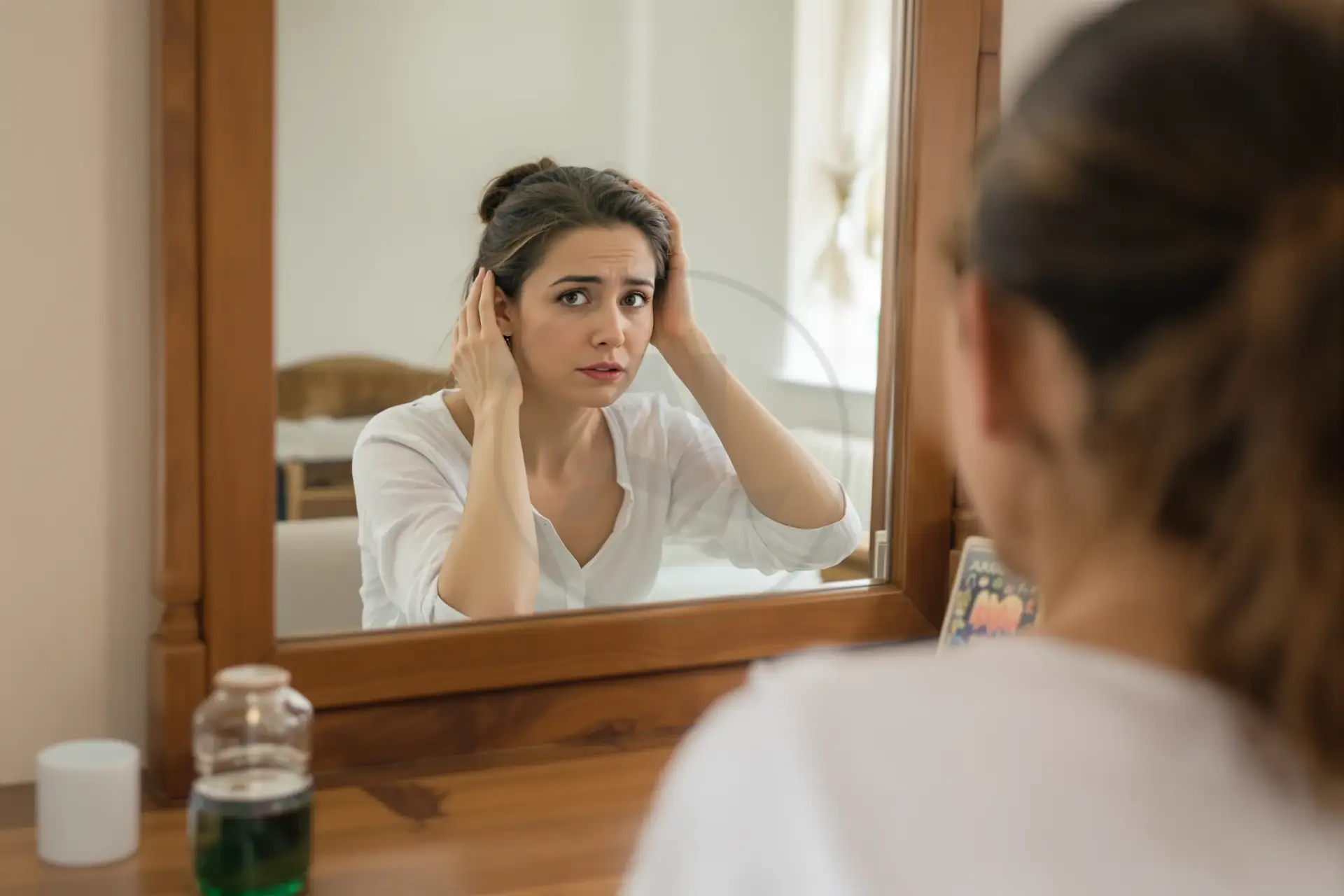 Woman examining her scalp and hairline in a vanity mirror with a worried look