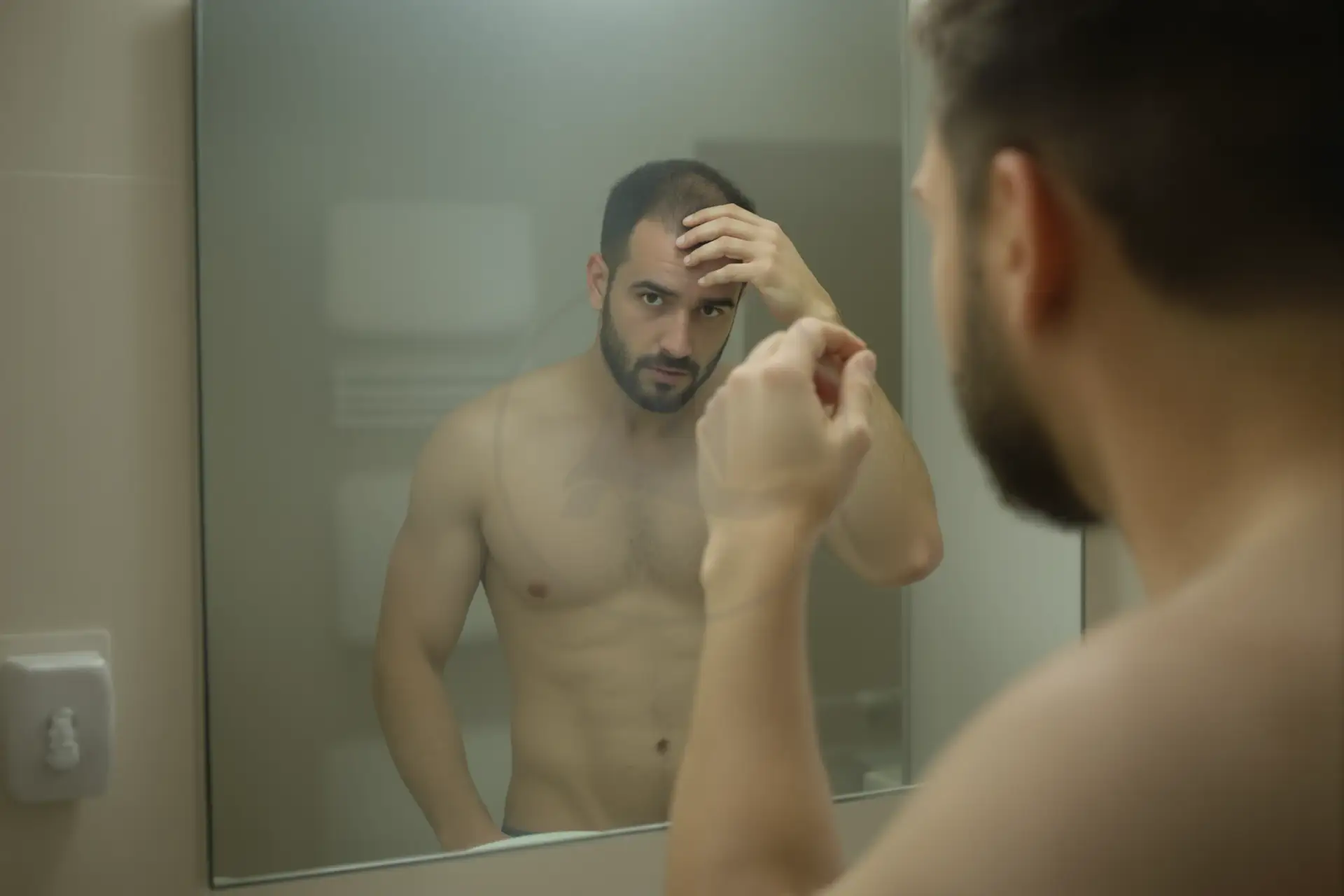 Shirtless bearded man examining his hairline in a bathroom mirror