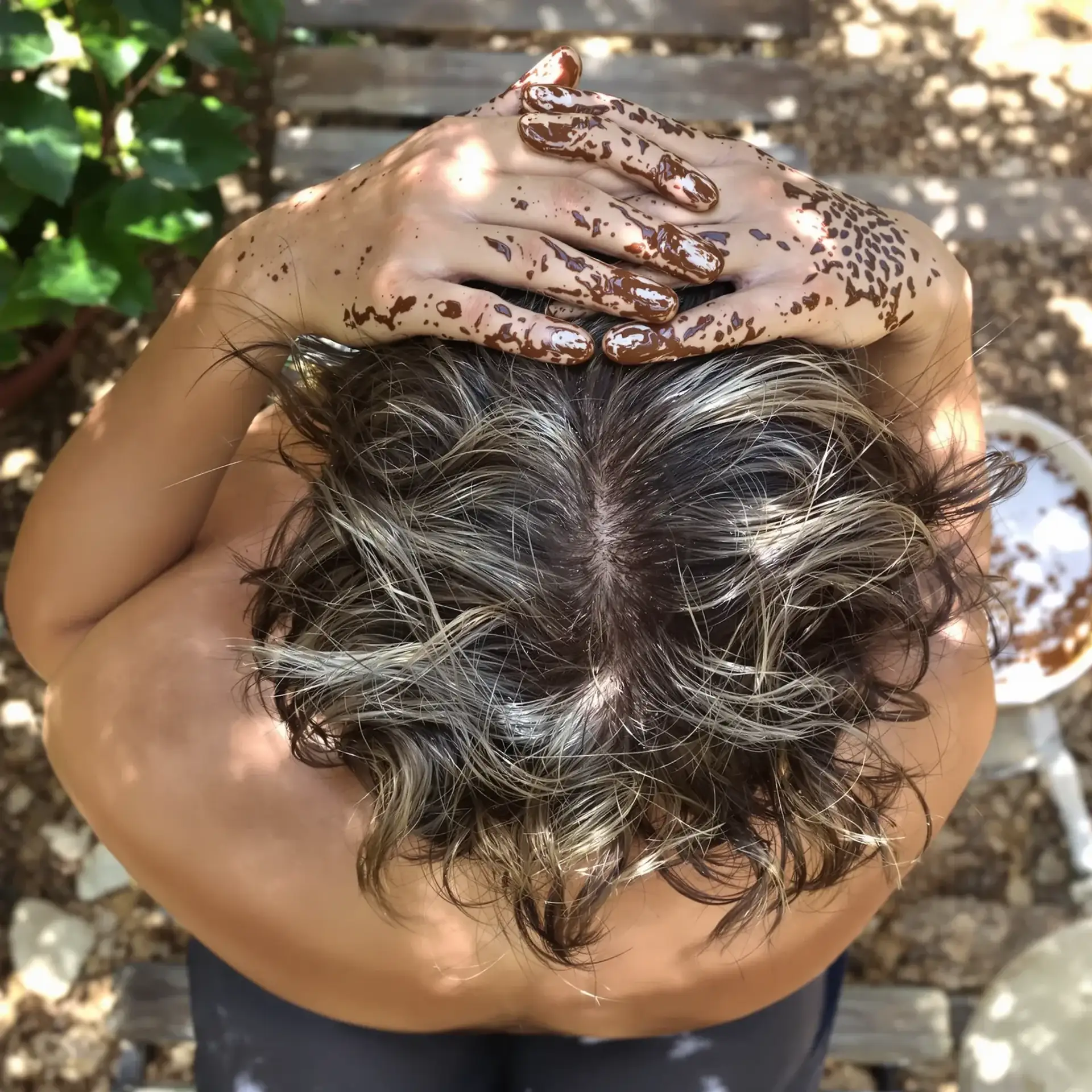 Overhead view of a person applying brown hair dye to their hair with dye-covered hands outdoors