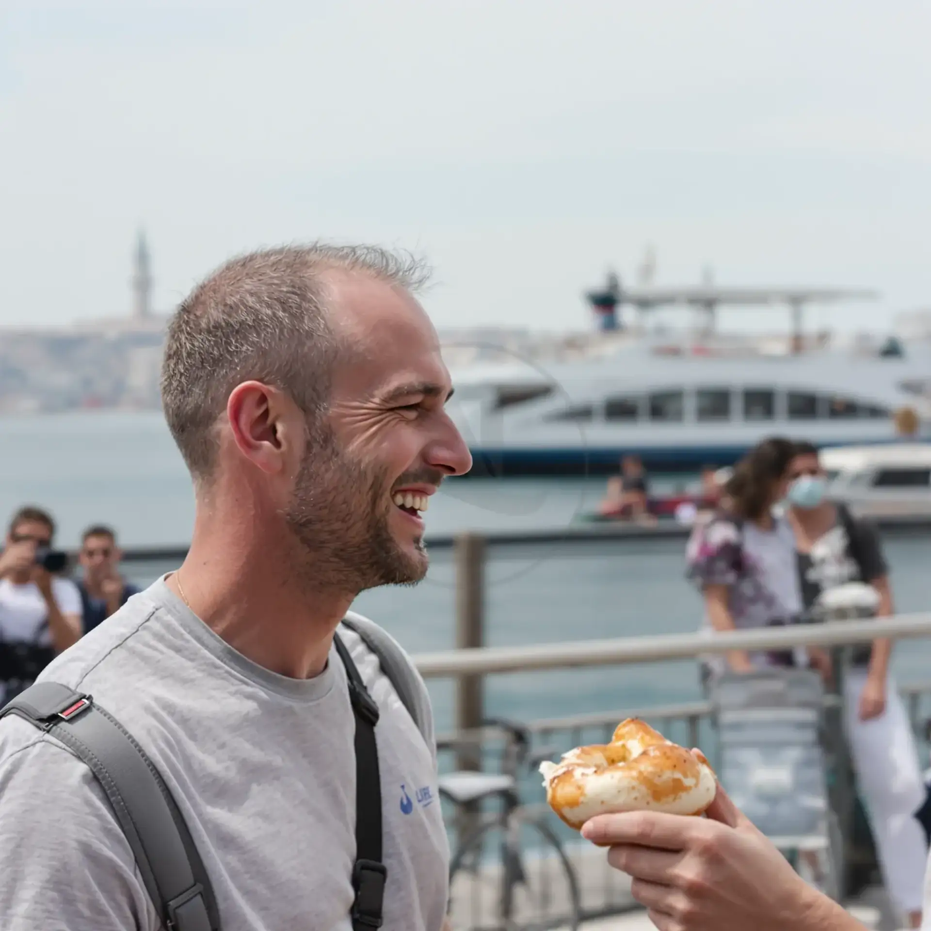 Smiling man with short hair by a waterfront eating a pastry with a ferry in the background