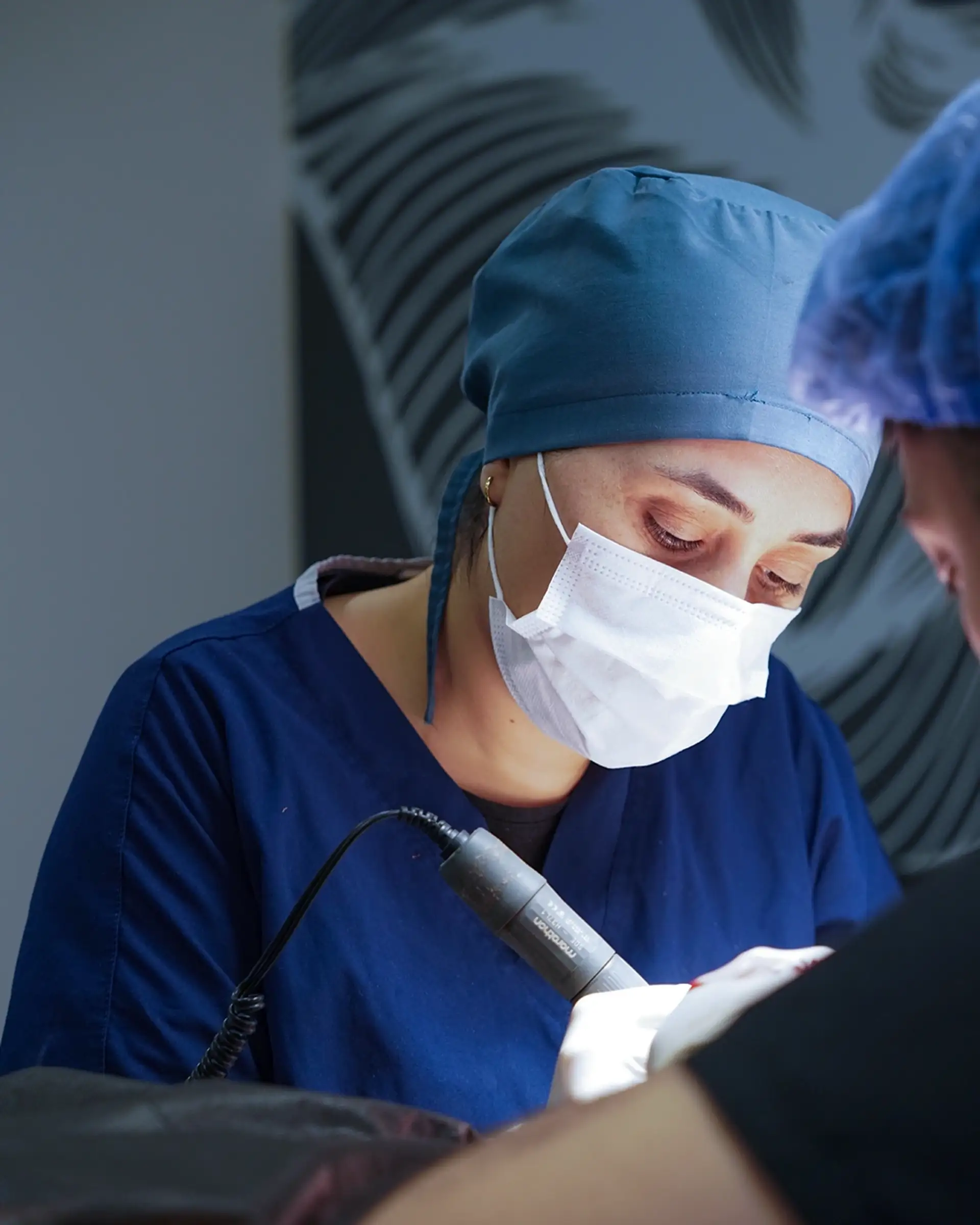 Istanbul Care team member in blue surgical scrubs, cap and face mask holding a corded motorised FUE handpiece while working over a patient in the treatment room.