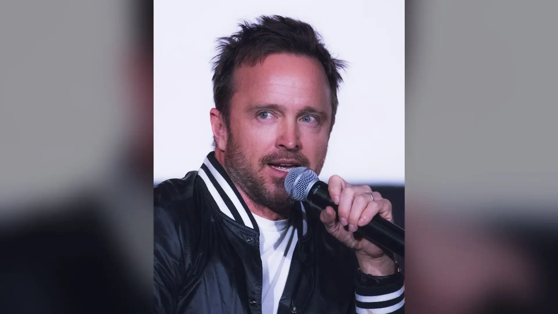 Aaron Paul speaking into a handheld microphone at a screening event, his short textured hair lit from above.