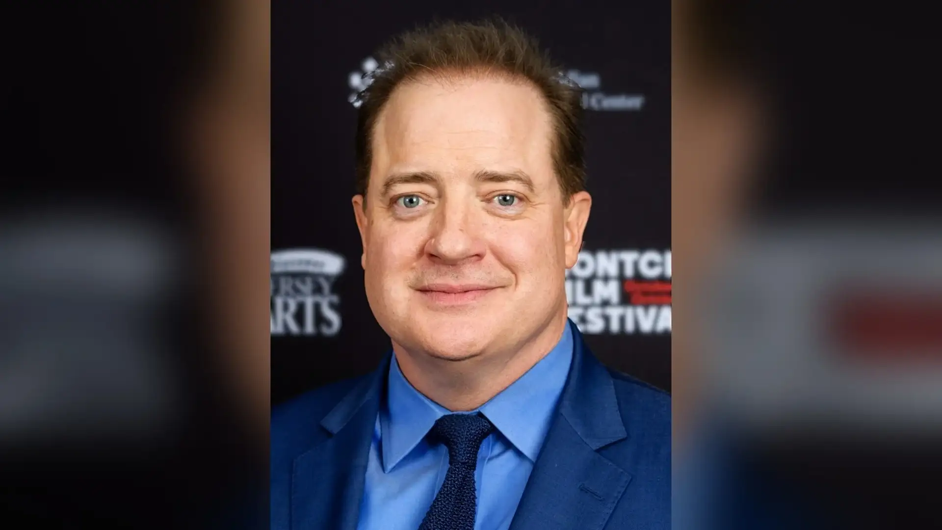 Brendan Fraser smiling in a blue suit at a film-festival press wall, his dark hair brushed back off the forehead.