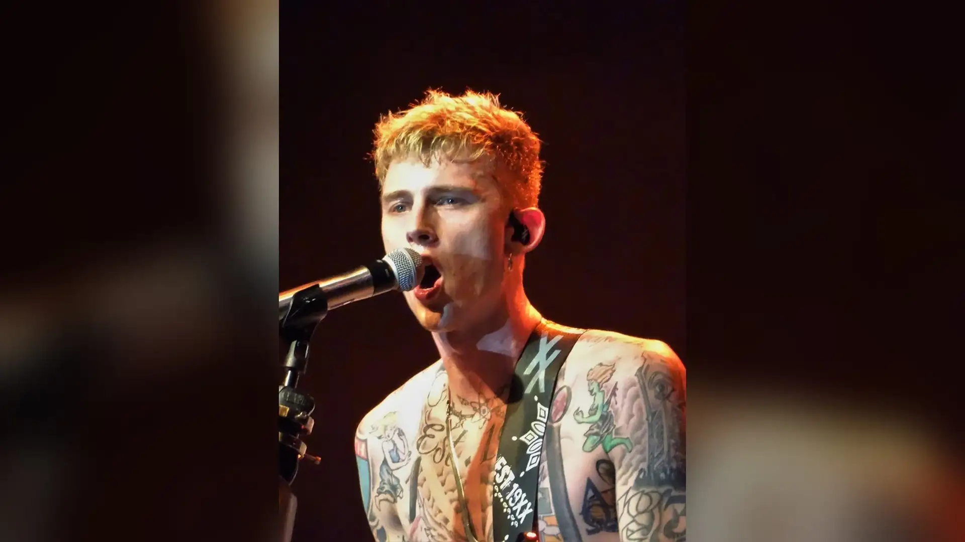 Machine Gun Kelly singing into a microphone under stage lighting, his short bleached hair and front hairline lit from above.