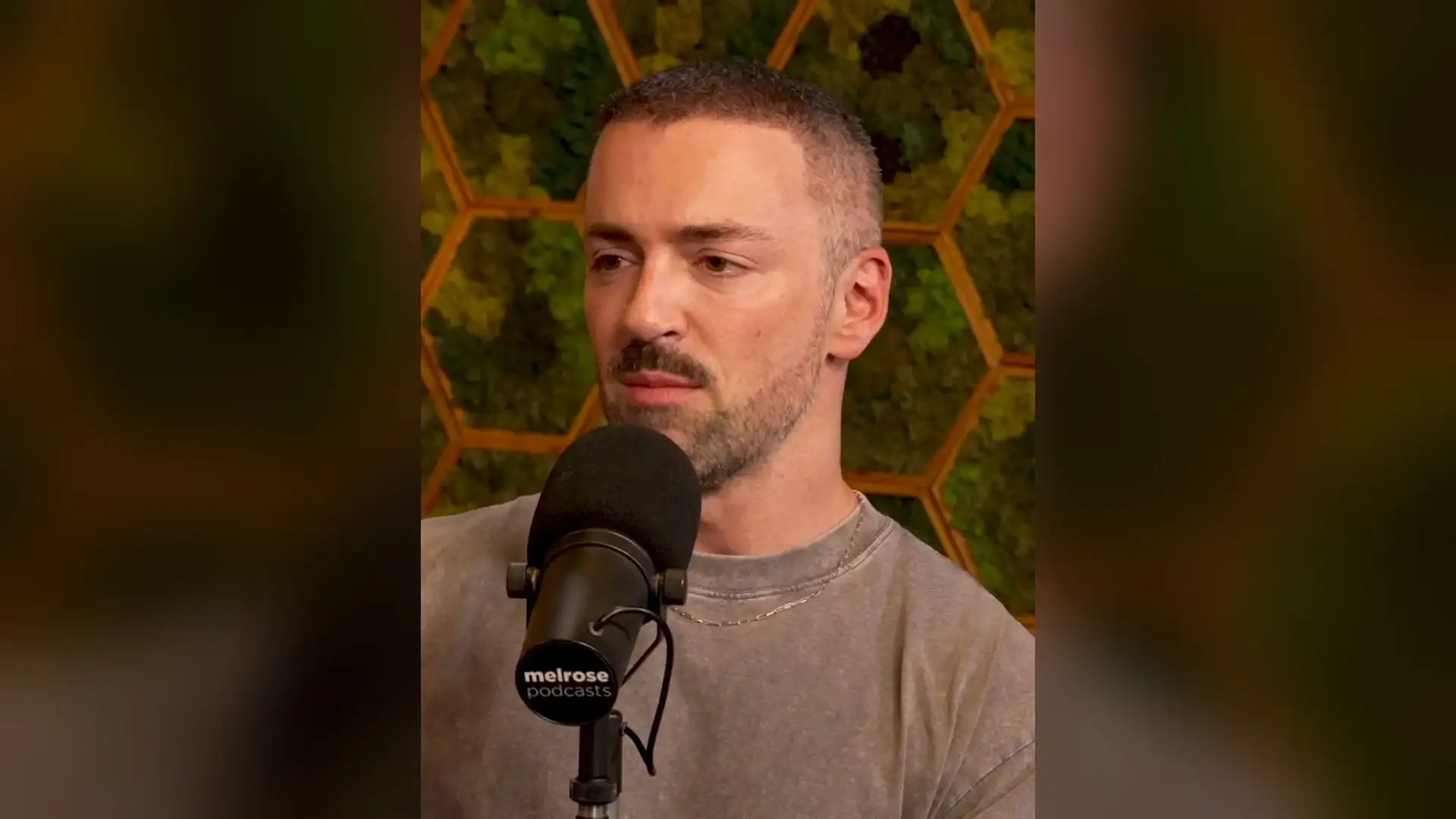 Matteo Lane sitting behind a podcast microphone in a beige T-shirt, his closely cropped hair and front hairline visible against a moss-panel wall.