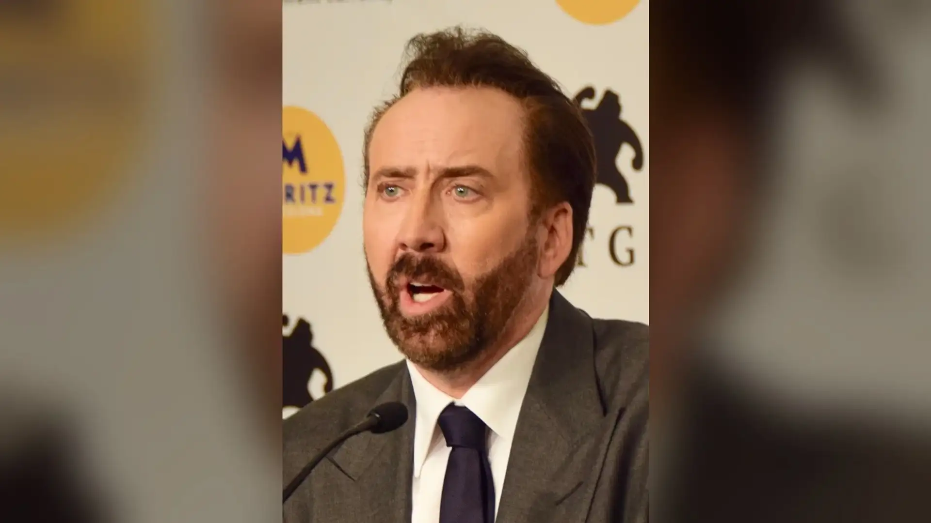 Nicolas Cage speaking at a press-conference microphone in a grey suit, seen from the front with his temples and hairline in view.