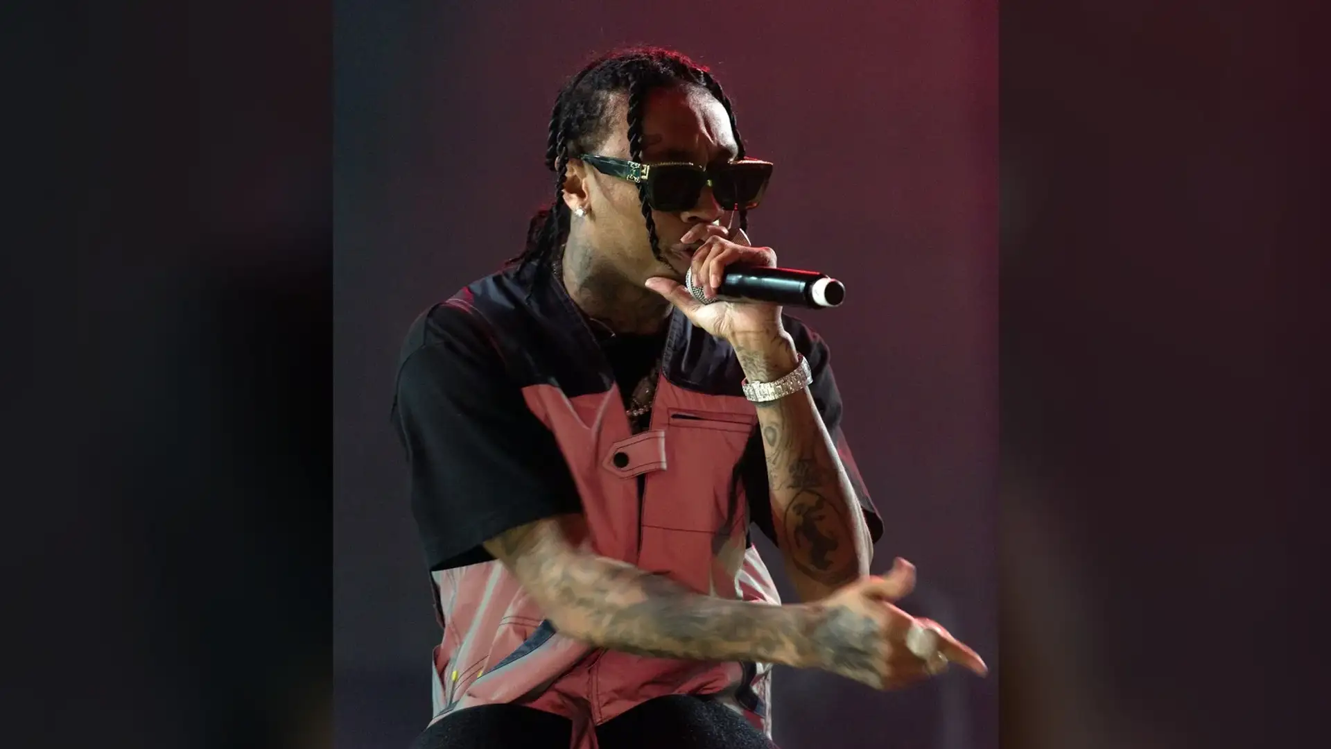 Tyga performing on stage in sunglasses, holding a microphone, with braided hair pulled back from the forehead.