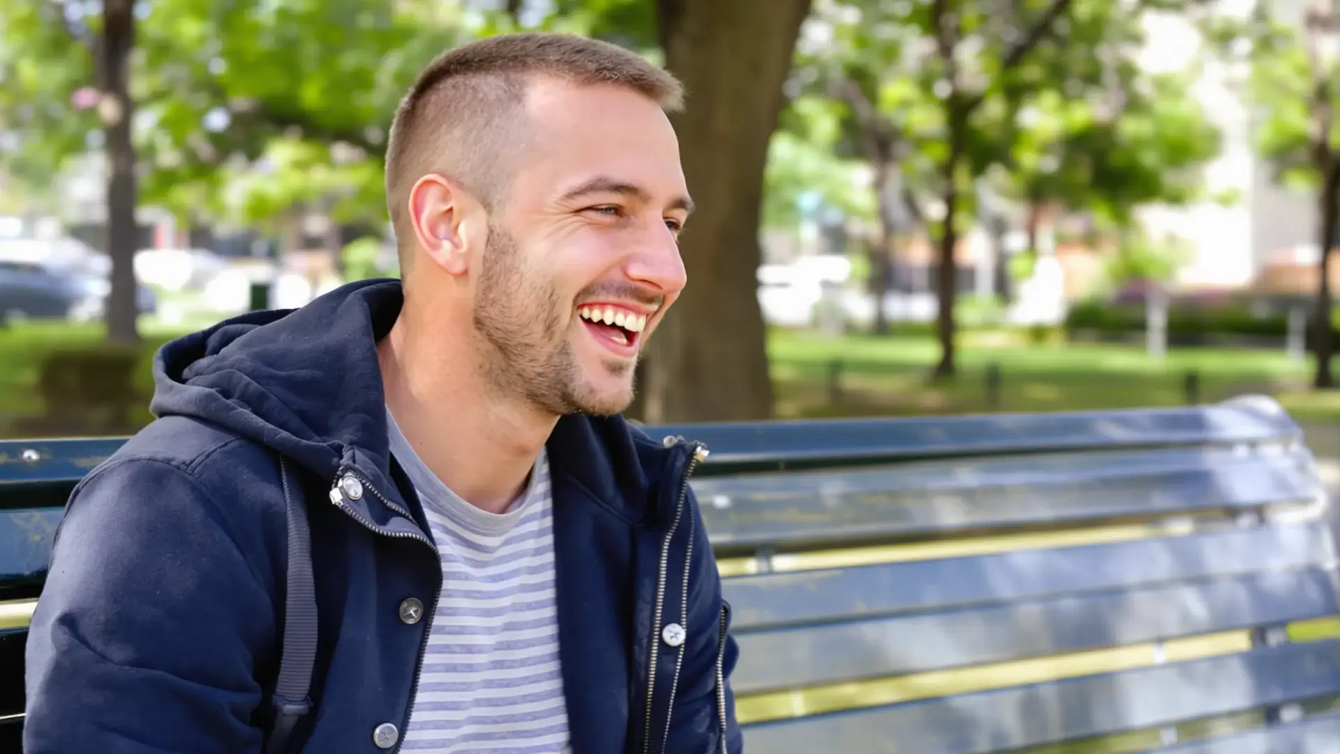 Young man with a full, straight hairline laughing on a park bench